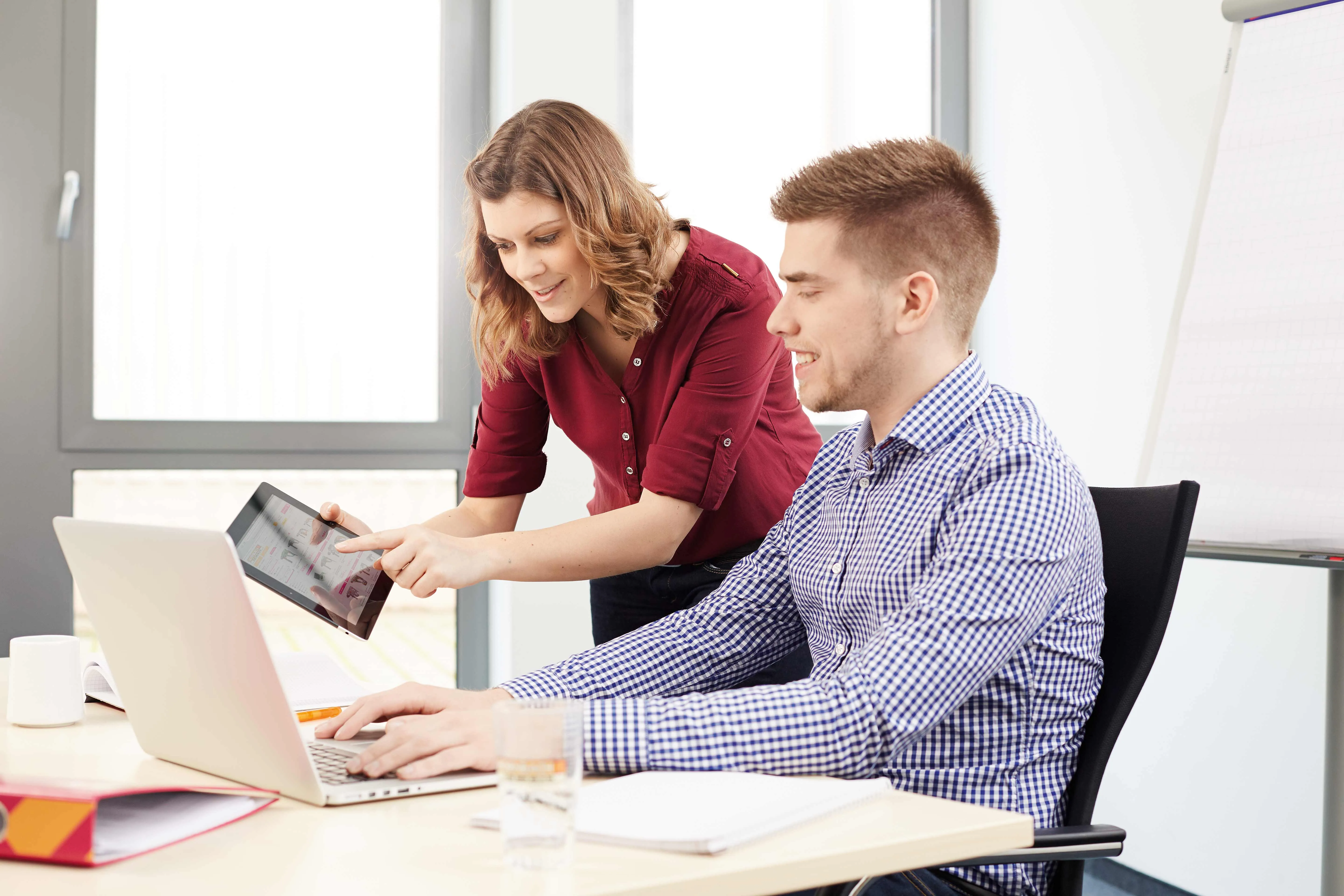 Zwei Kollegen in einem modernen Büro, die an einem Laptop zusammenarbeiten. Eine Frau zeigt einem Mann etwas auf einem Tablet.