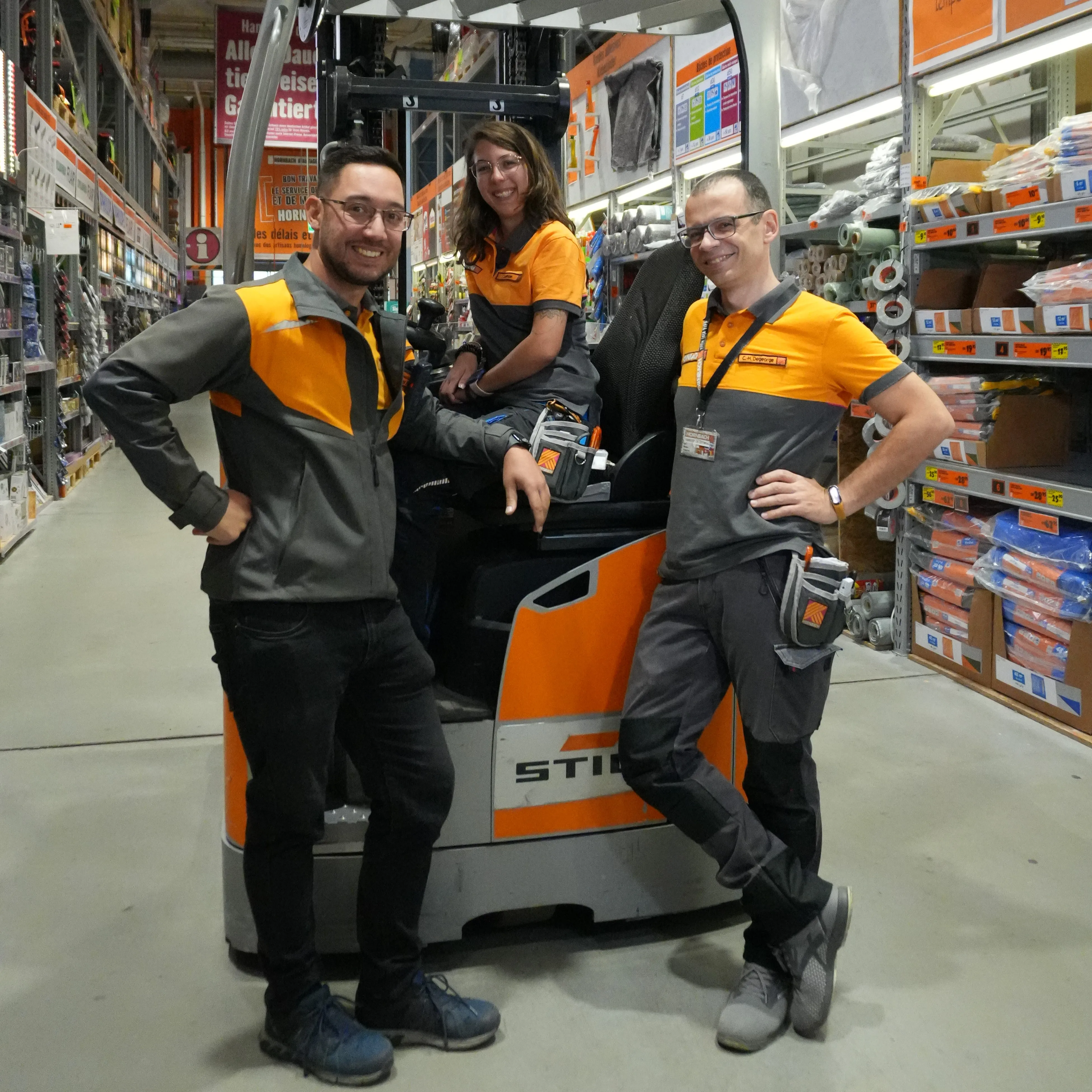 Trois collègues souriants de HORNBACH dans un magasin, posant près d'un chariot élévateur.