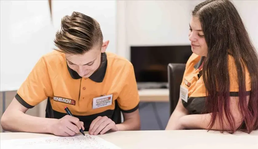 Deux employés de HORNBACH en uniforme discutent et prennent des notes ensemble dans un environnement de bureau.