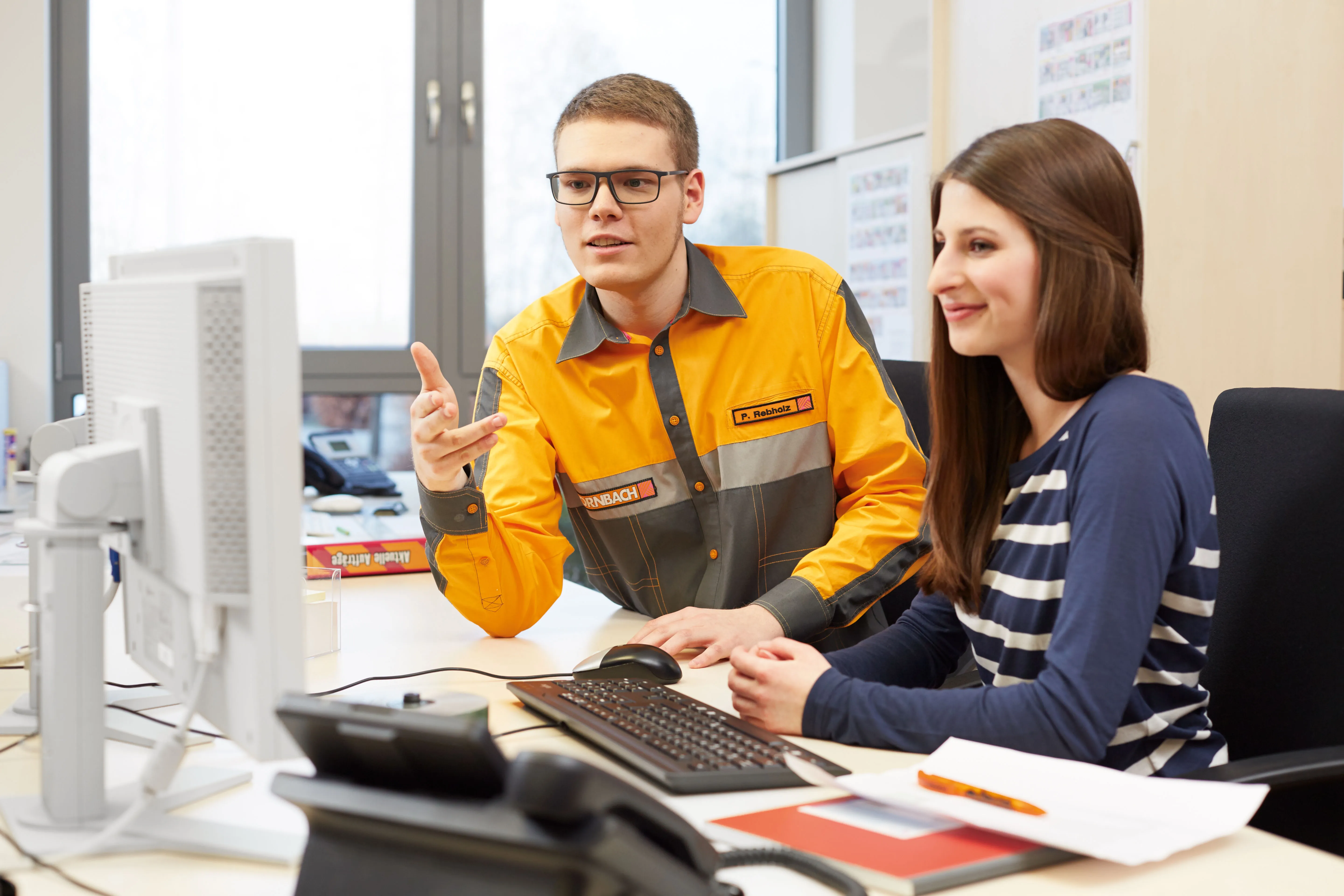 Mitarbeiter im HORNBACH-Logo erklärt einer Kollegin am Computer einen Prozess im Büro.
