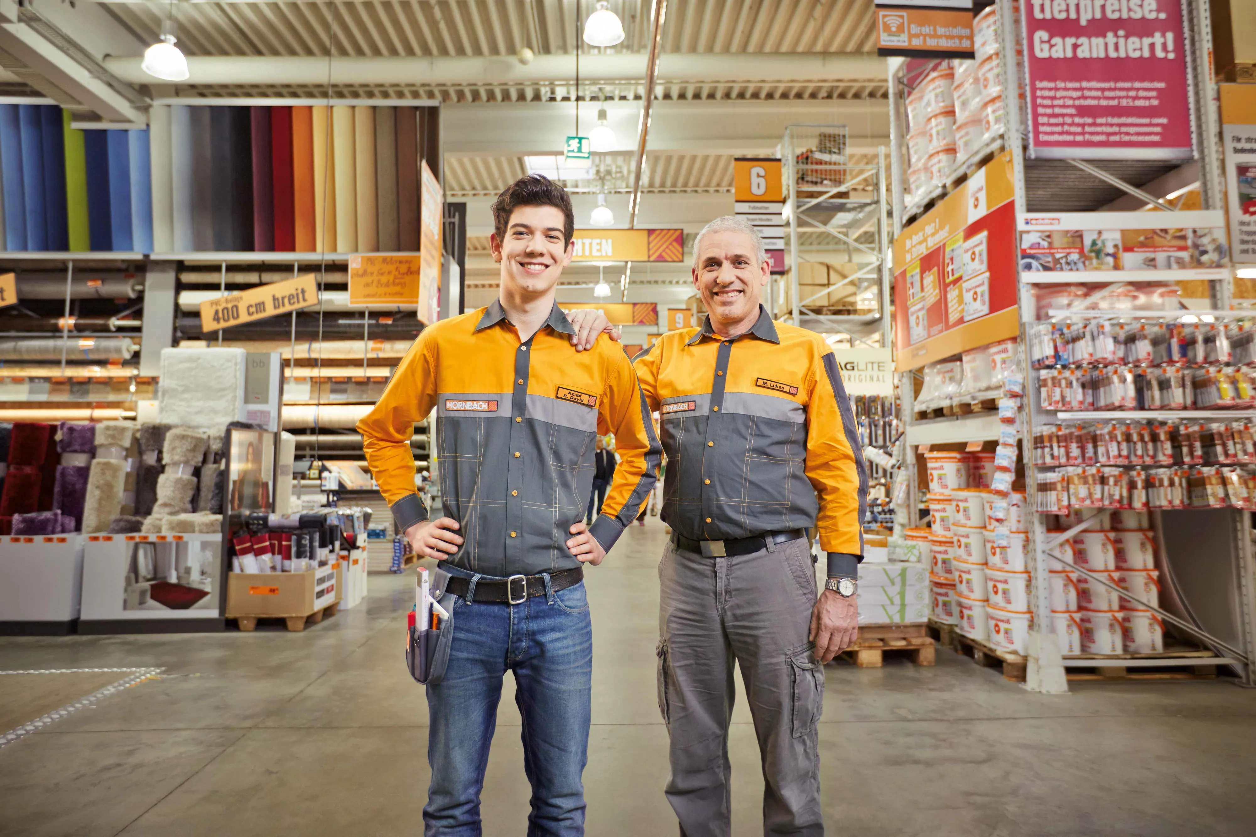 Zwei lächelnde HORNBACH-Mitarbeiter in Arbeitskleidung stehen in einem Baumarktgang vor Regalen mit Heimwerkerbedarf.