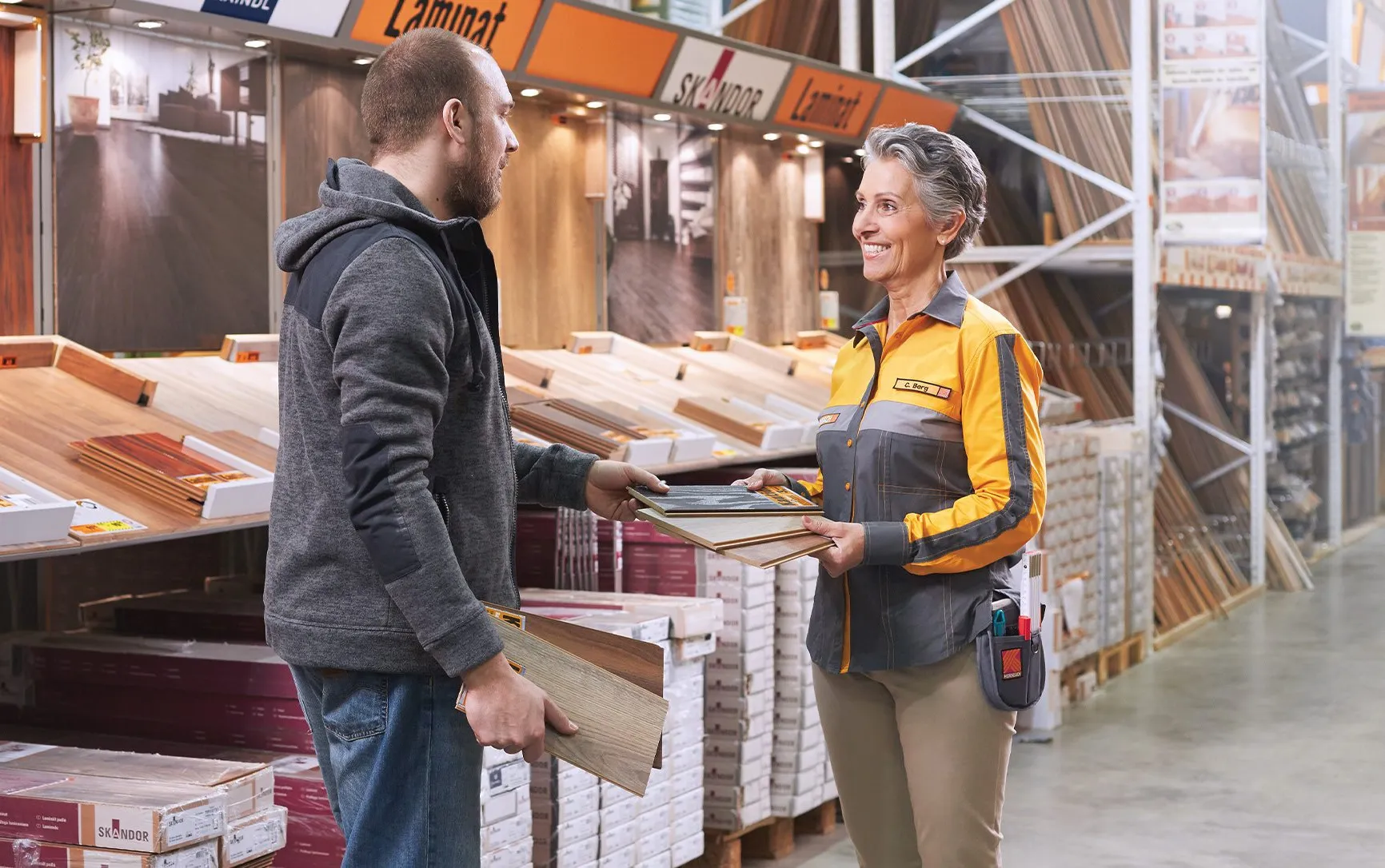 Un client discute avec une employée de HORNBACH, tenant des échantillons de parquet dans le rayon revêtements de sol.