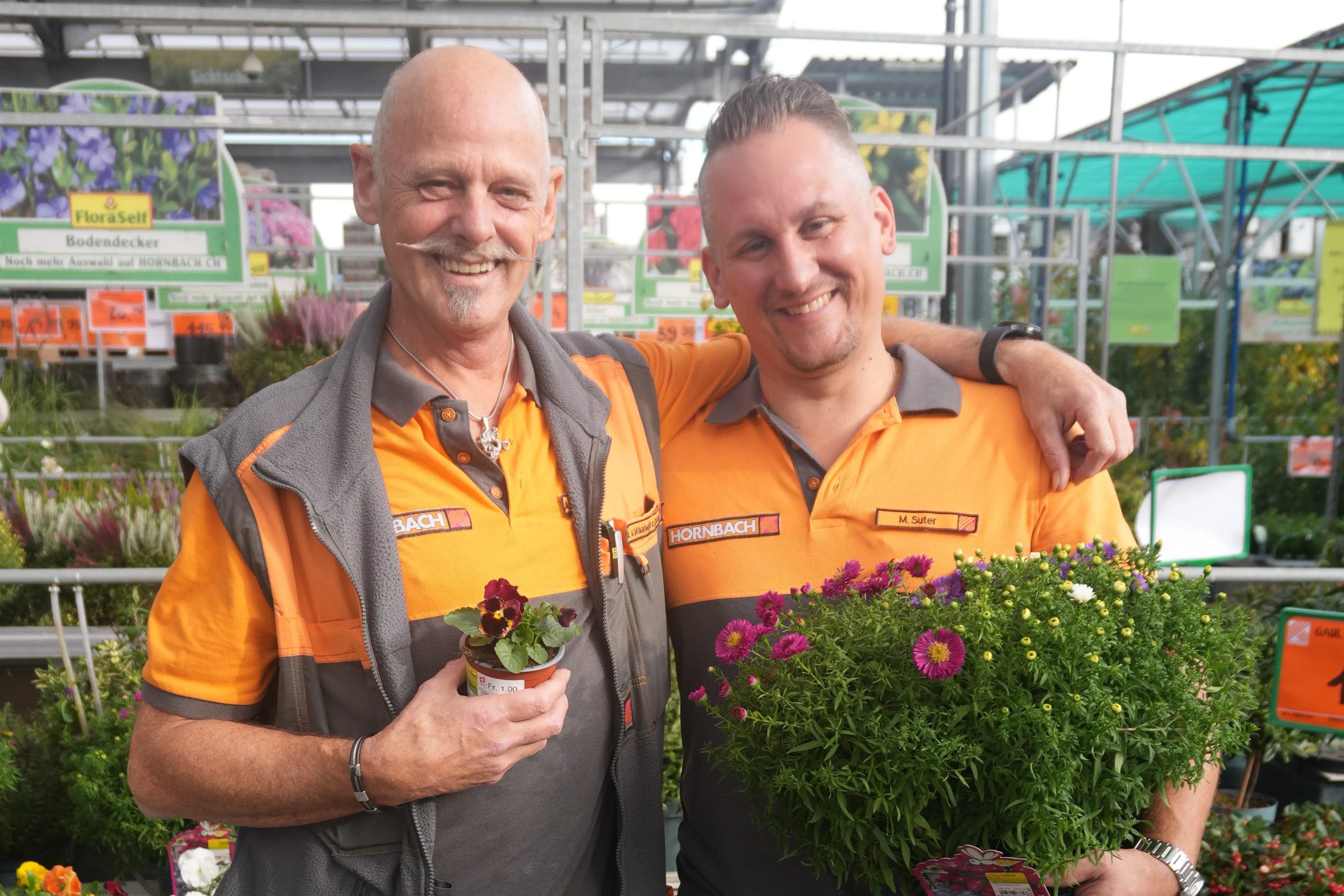 Zwei lächelnde HORNBACH-Mitarbeiter in der Gartenabteilung, einer hält eine Topfblume, der andere einen Blumentopf.