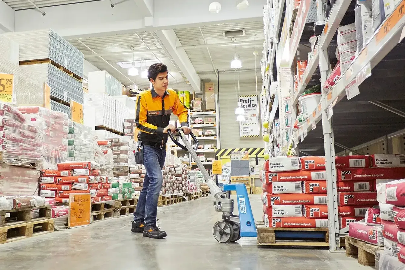 Un employé de HORNBACH Baumarkt AG déplace des palettes dans un magasin de bricolage, portant un uniforme orange et noir.