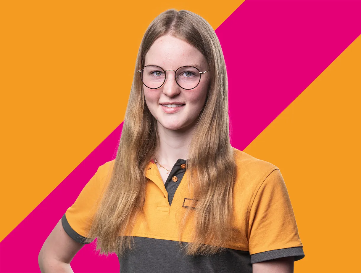 Eine Mitarbeiterin lächelt in die Kamera vor einem farbenfrohen Hintergrund aus Orange und Pink.