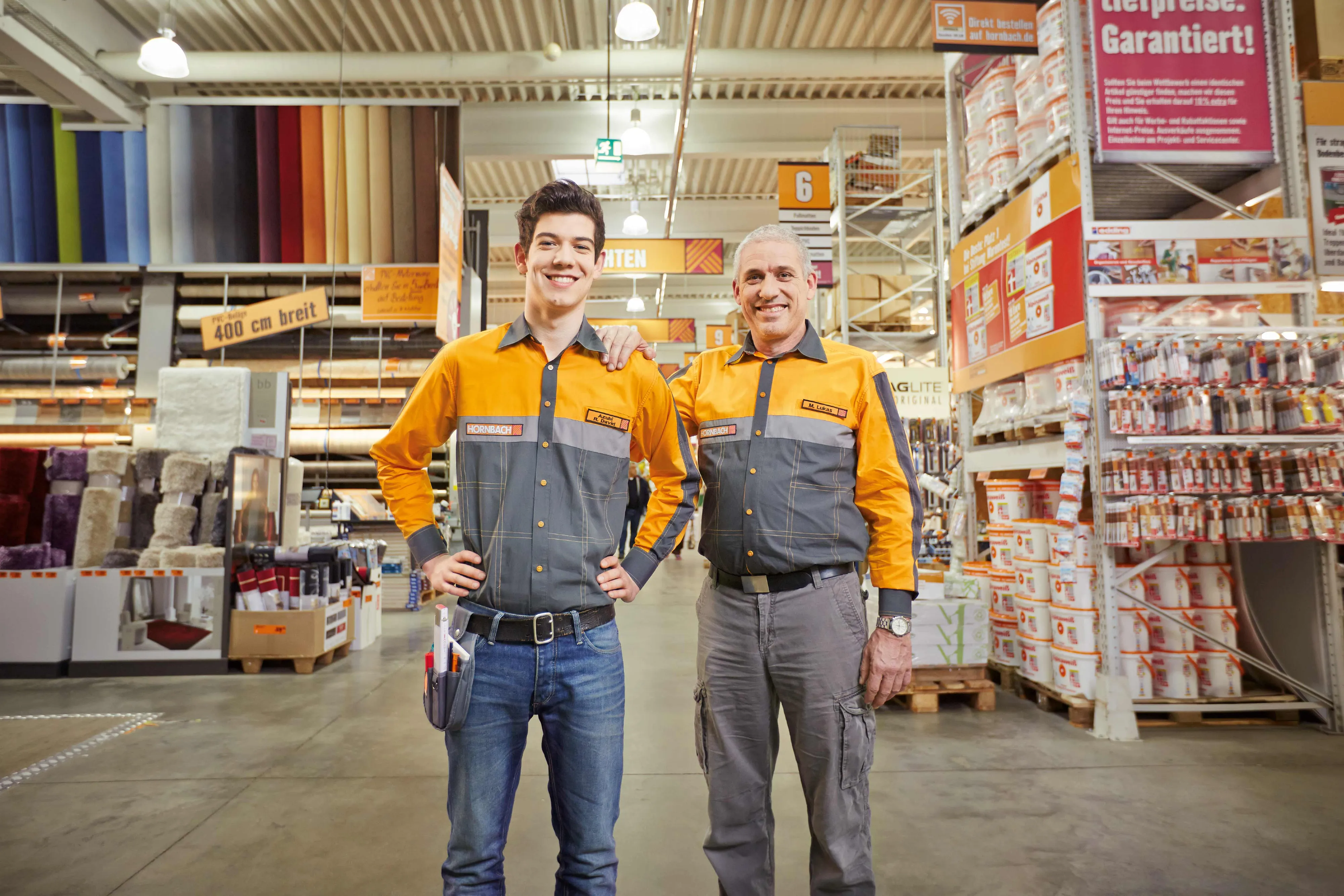 Deux employés souriants en uniforme HORNBACH posent dans un magasin de bricolage, entourés de matériaux et outils.