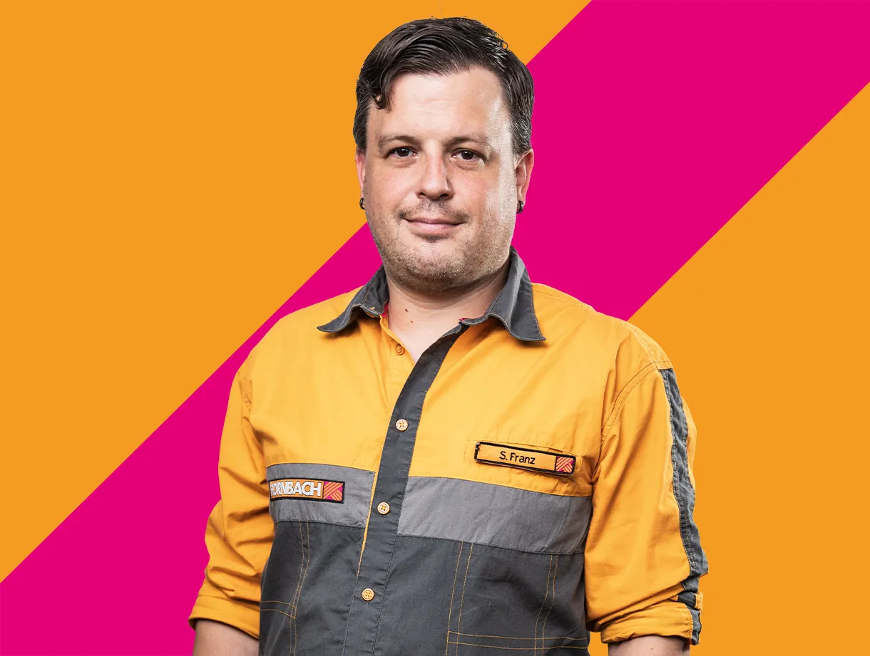 Eine Mitarbeiterin von HORNBACH in orange-grauer Arbeitskleidung vor einem farbenfrohen Hintergrund.