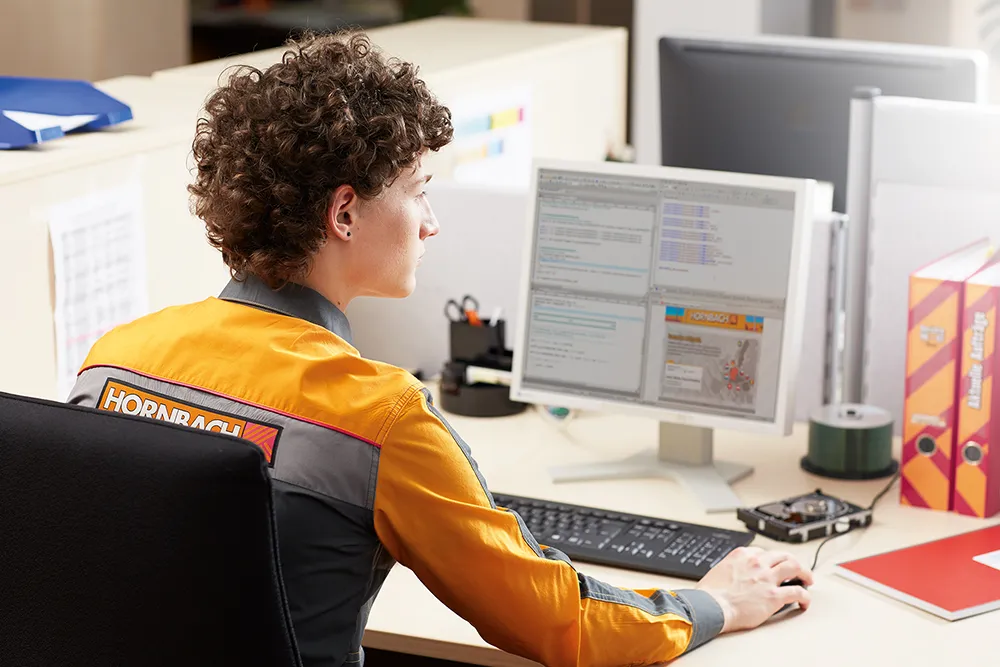 Ein Mitarbeiter von HORNBACH arbeitet am Computer im Büro. Du siehst das HORNBACH-Logo auf seiner Arbeitskleidung.