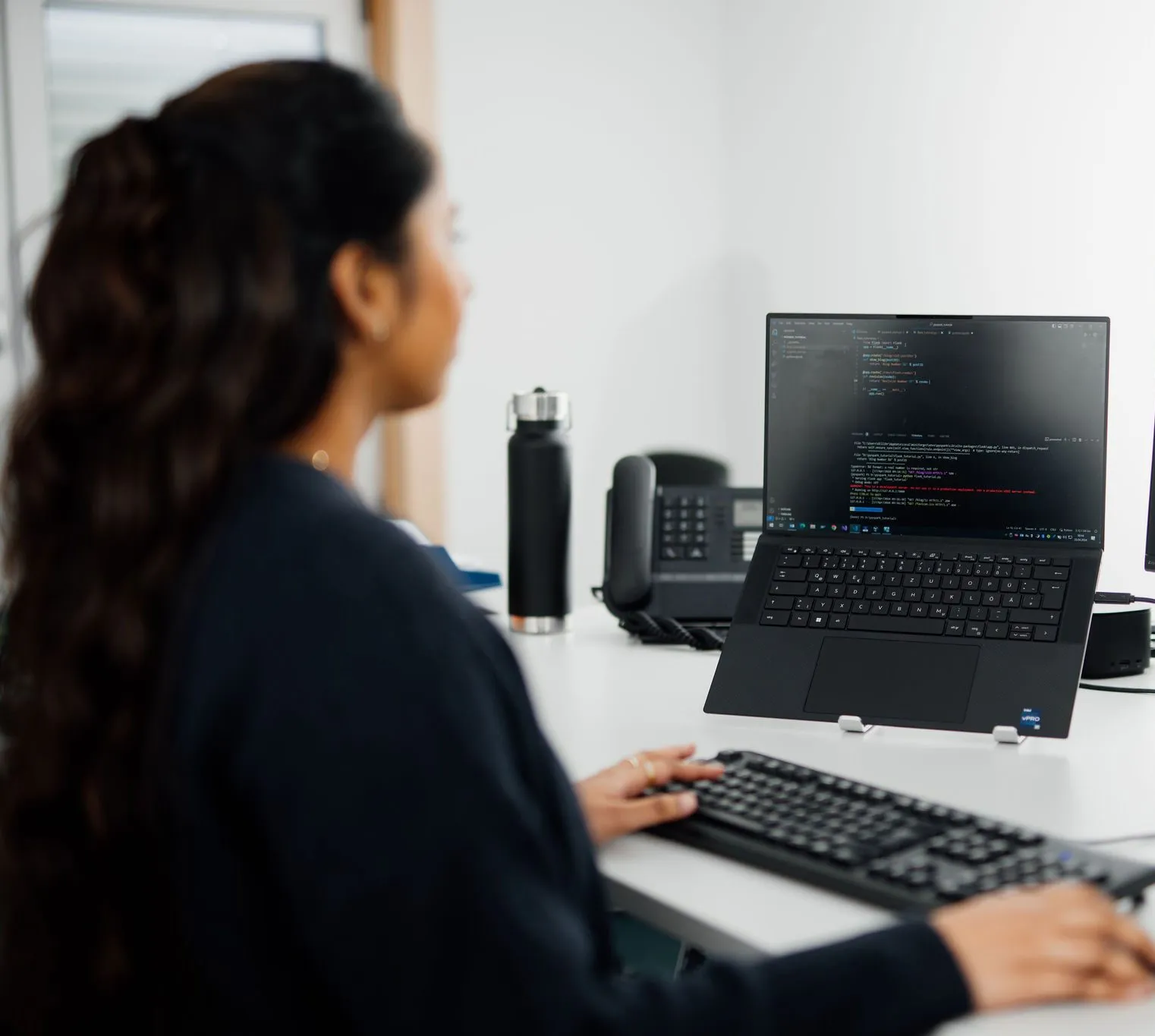 Eine Frau mit Headset arbeitet lächelnd an einem Laptop in einem modernen Büro.
