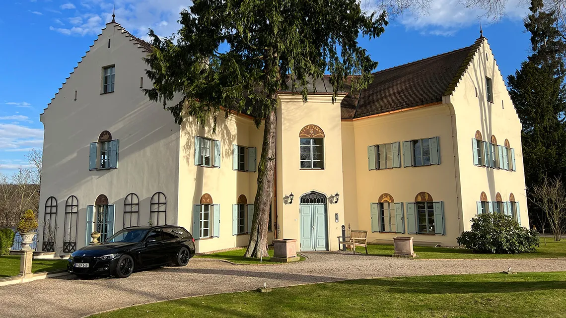 Helles Firmengebäude von impeak GmbH mit gepflegtem Garten und schwarzem Auto vor dem Eingang, bei sonnigem Wetter.