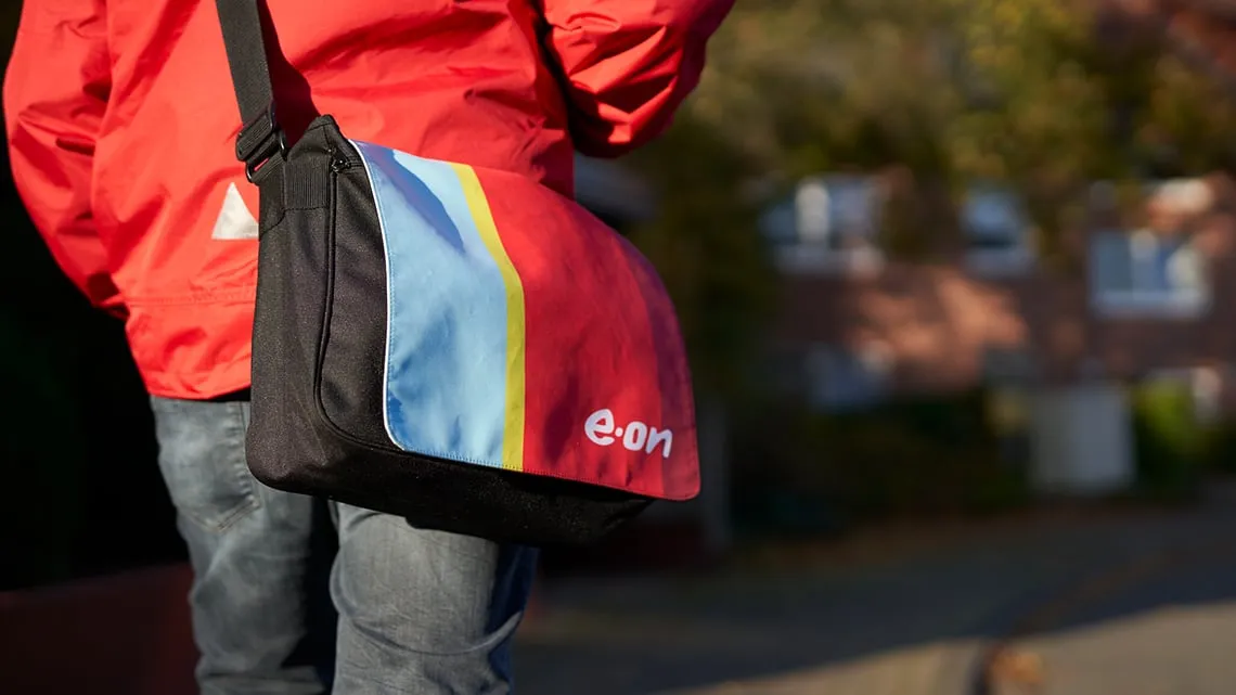 Du trägst eine bunte Umhängetasche mit e.on-Logo und roter Jacke, bereit für deinen Tag im Außendienst.