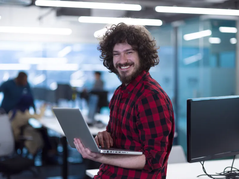 Junger Mann im modernen Büro hält lächelnd einen Laptop – starte Deine Karriere bei Interhyp AG im Finanzbereich.
