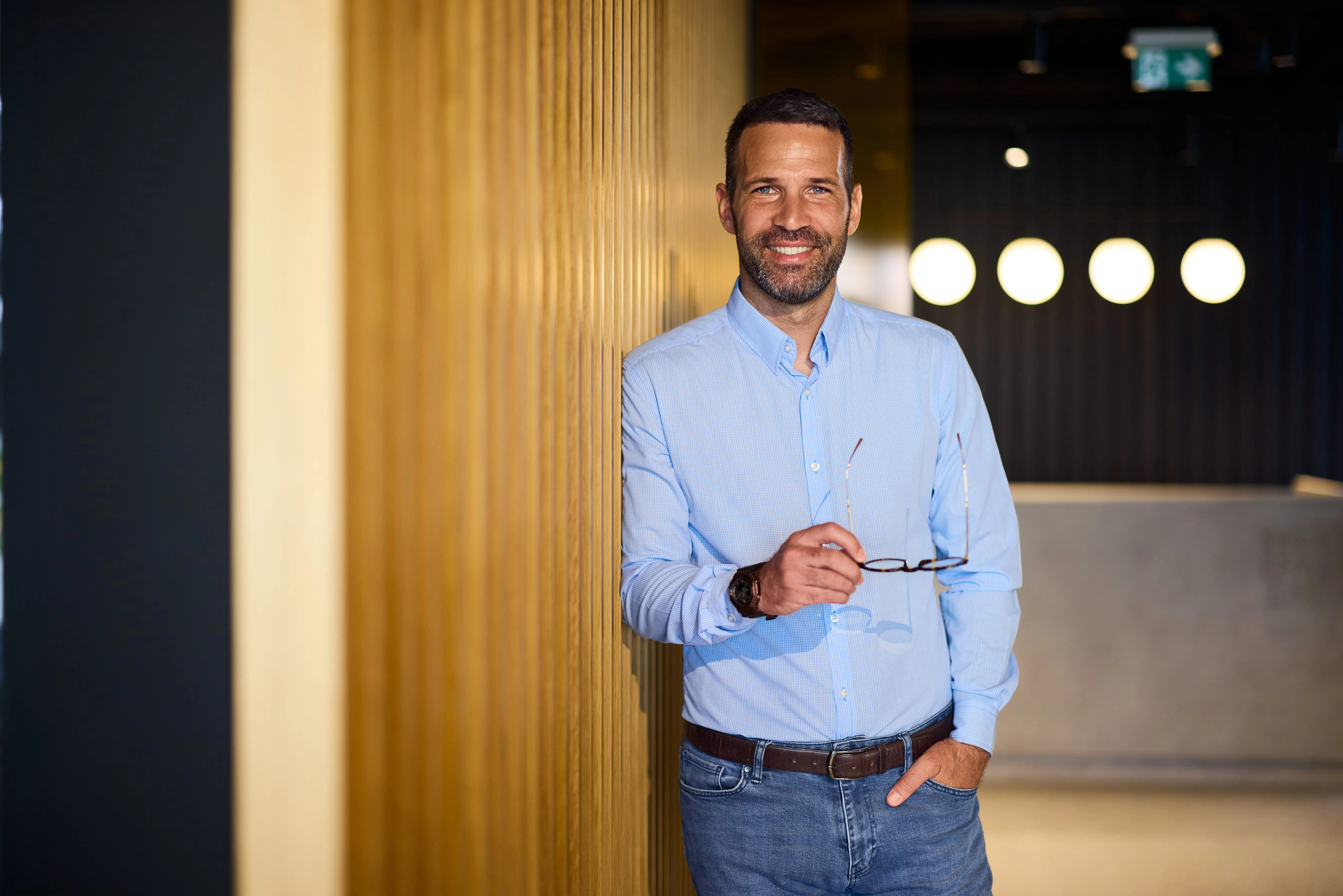 Ein Mann im Büro von Interhyp AG, lässig an eine Holzverkleidung gelehnt, hält eine Brille und lächelt in die Kamera.