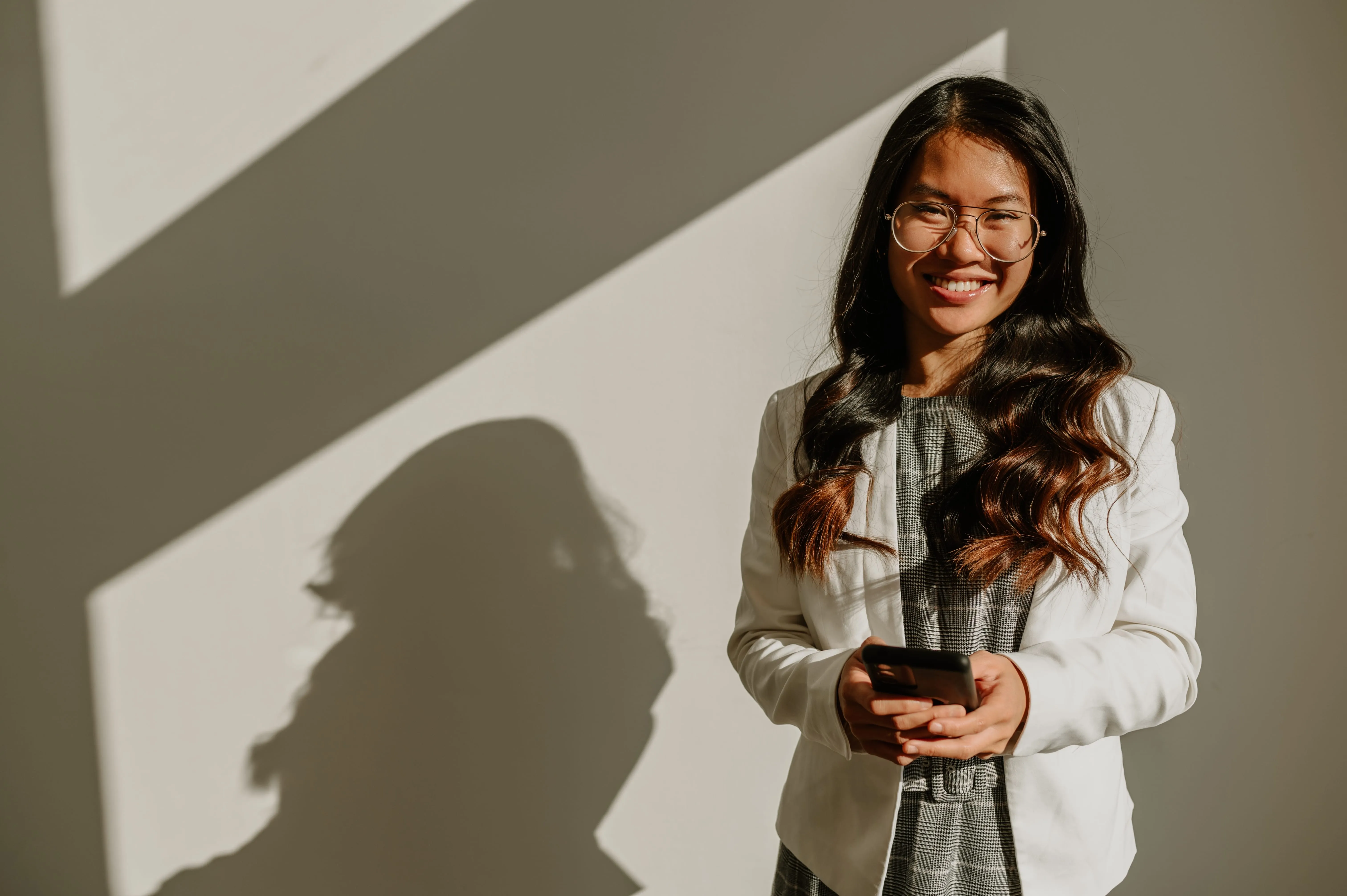 Eine lächelnde Frau im Business-Outfit steht mit einem Smartphone in der Hand vor einer Wand.
