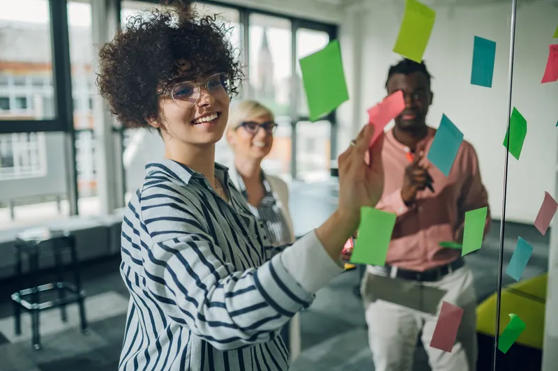 Drei junge Menschen arbeiten gemeinsam an einer Glaswand mit bunten Post-its in einem modernen Büro.