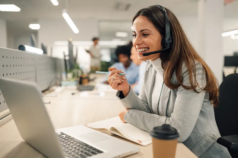 Junge Frau im Büro mit Headset, lächelt während eines Videocalls und macht sich Notizen am Laptop.