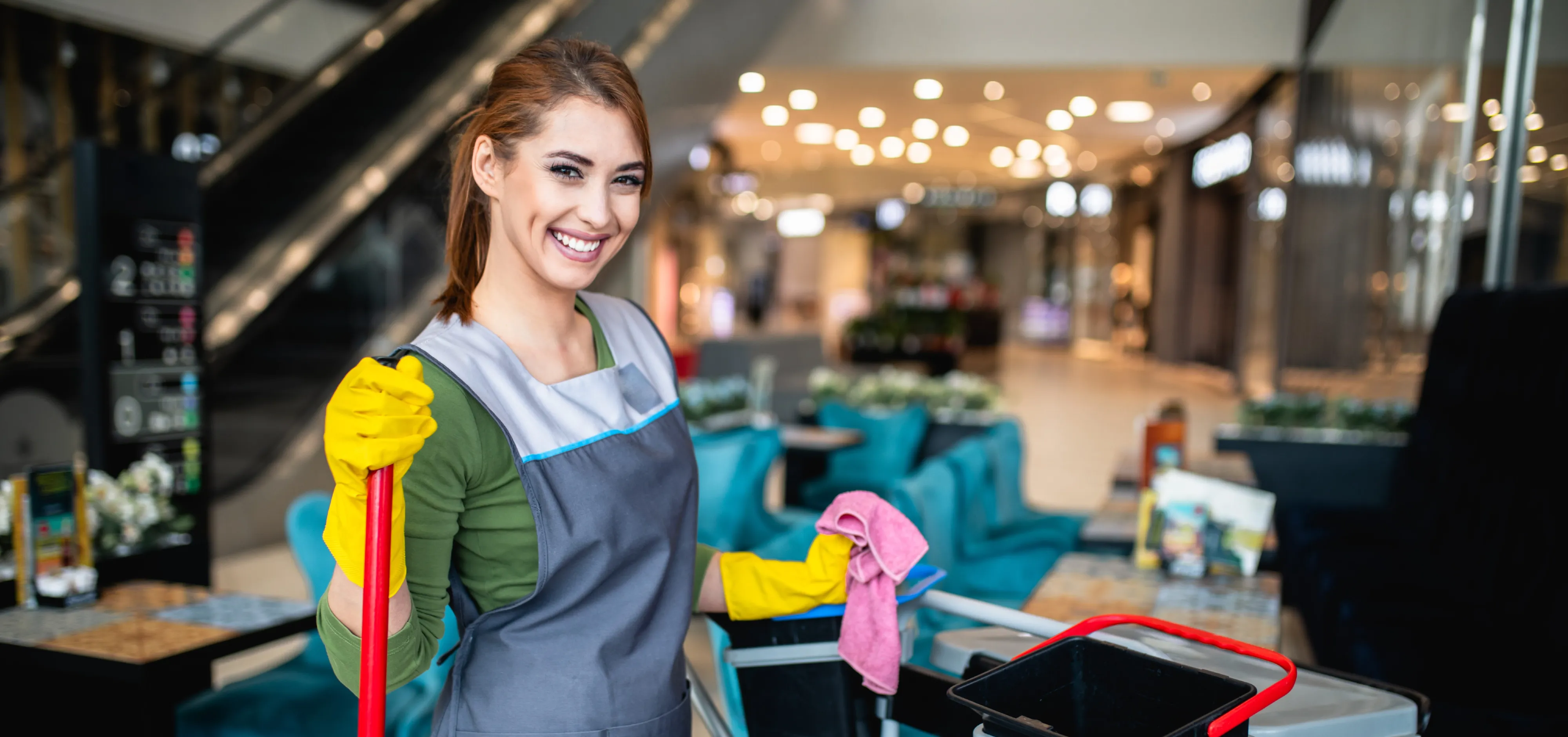 Eine lächelnde Reinigungskraft in Arbeitskleidung mit gelben Handschuhen hält einen Wischmopp in einem modernen Gebäude.