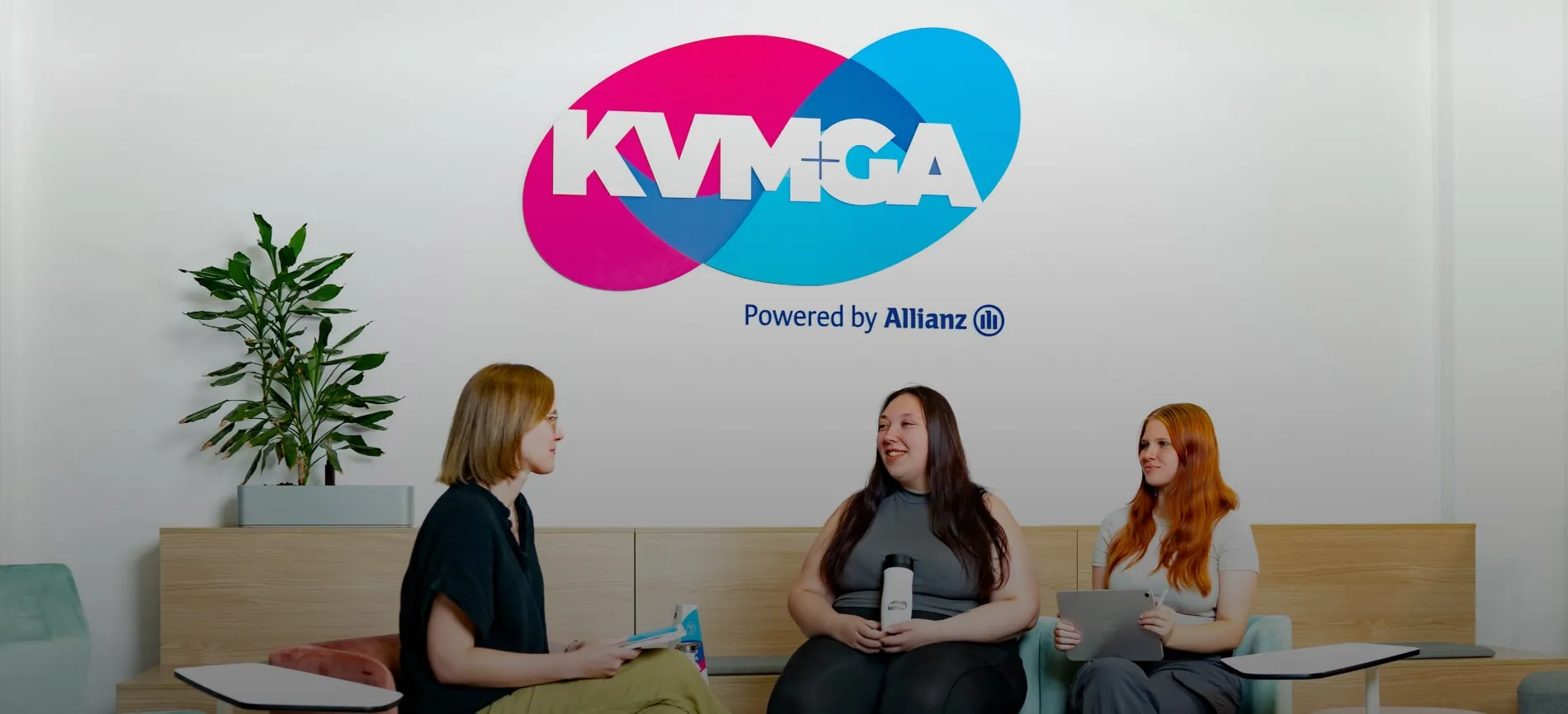 Drei junge Frauen unterhalten sich entspannt im modernen Büro von KVM ServicePlus, Logo im Hintergrund sichtbar.
