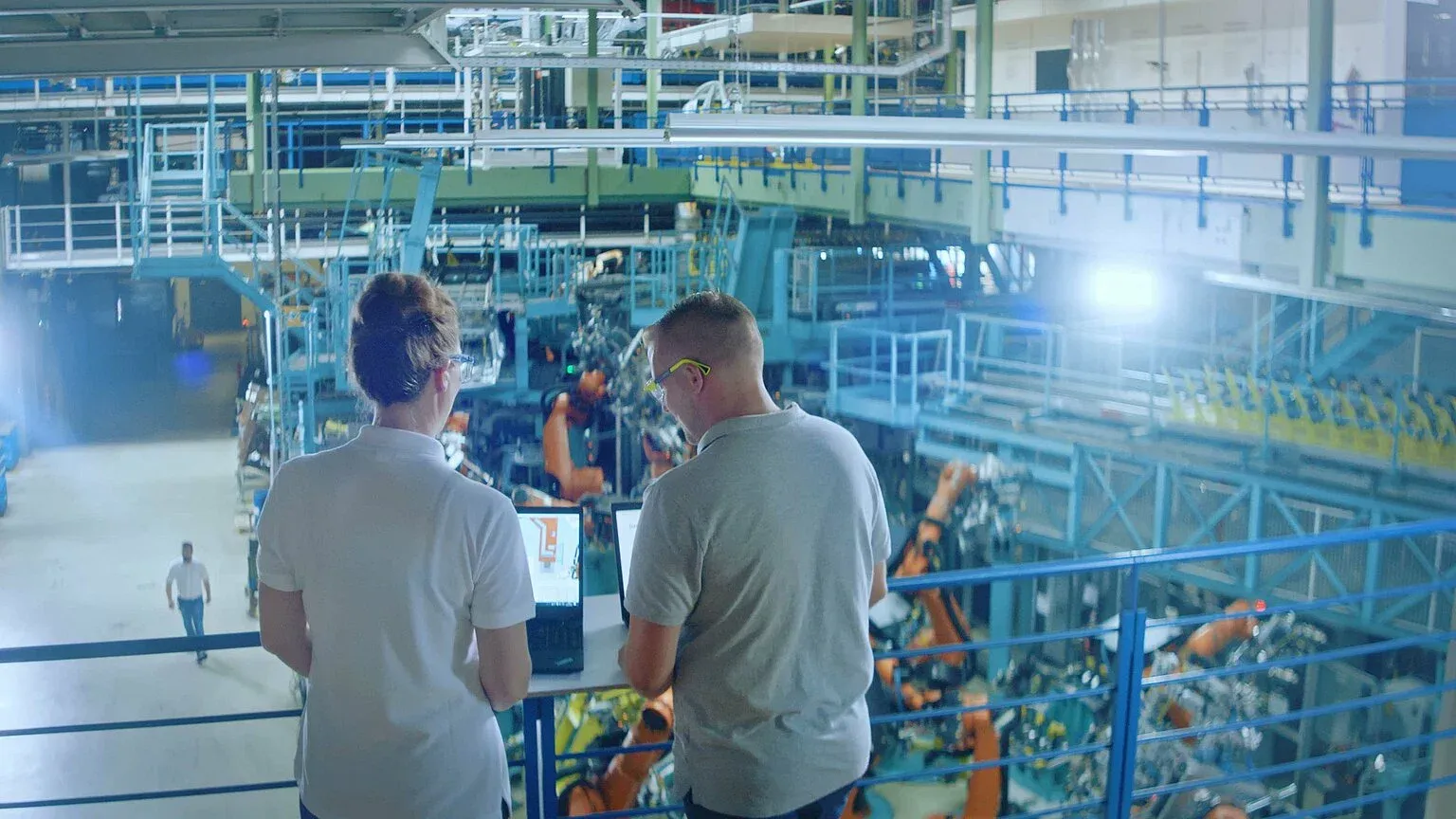 Zwei Mitarbeitende blicken auf eine moderne Produktionshalle mit Robotern und arbeiten an Laptops.