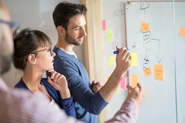 Drei Kollegen arbeiten gemeinsam an einem Whiteboard mit Skizzen und Post-its und besprechen Projektideen.