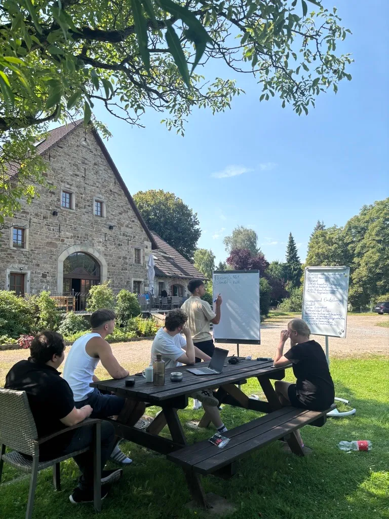 Vier junge Menschen sitzen an einem Picknicktisch im Grünen und brainstormen gemeinsam bei einem Team-Workshop.
