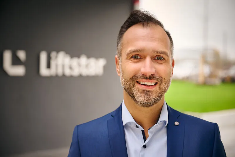 Ein lächelnder Mann im Anzug steht vor dem Liftstar-Logo und begrüßt Dich auf der Karriereseite im Gesundheitsbereich.
