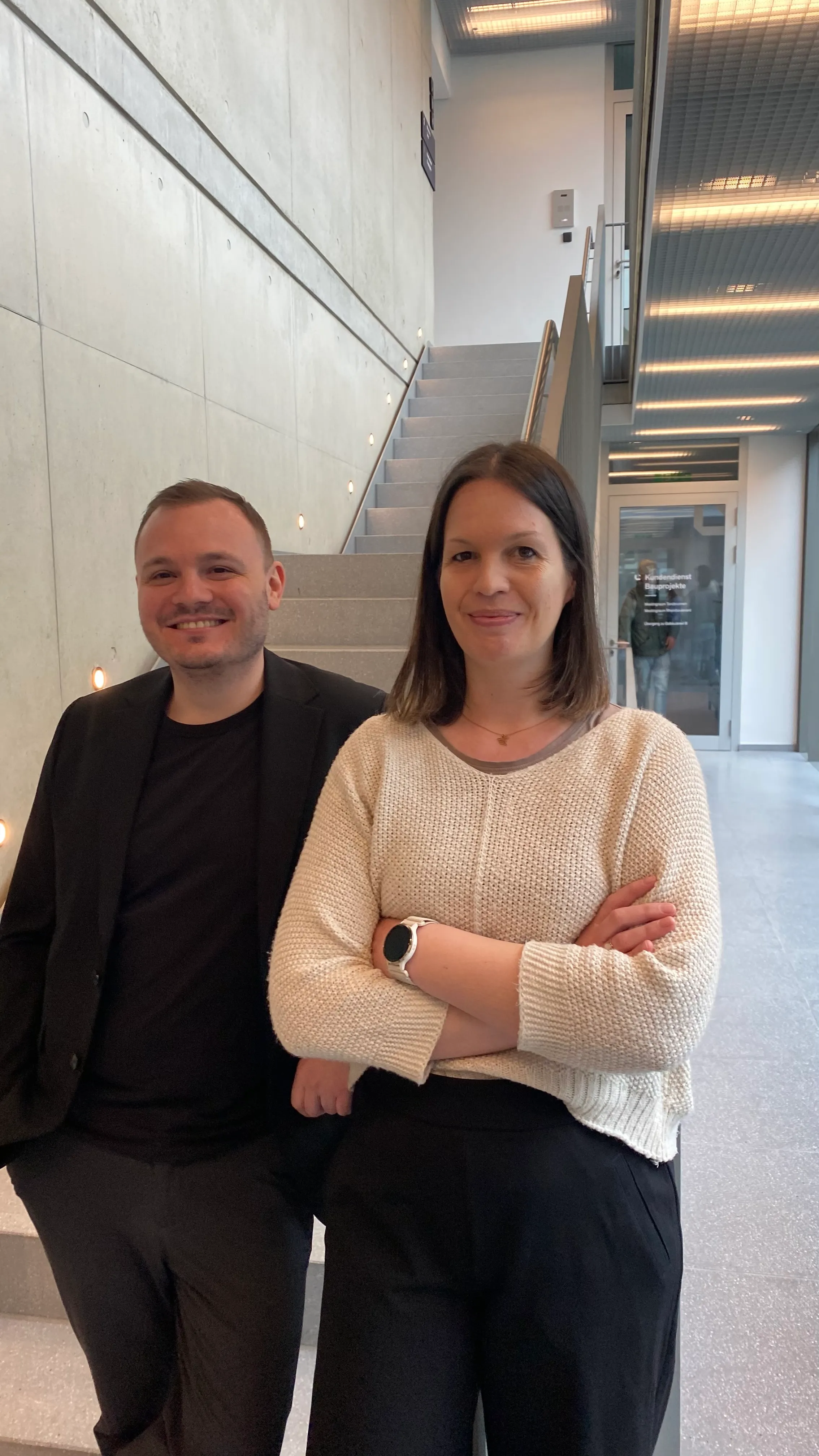Zwei freundliche Mitarbeitende von Liftstar GmbH stehen im modernen Bürogebäude und lächeln in die Kamera.