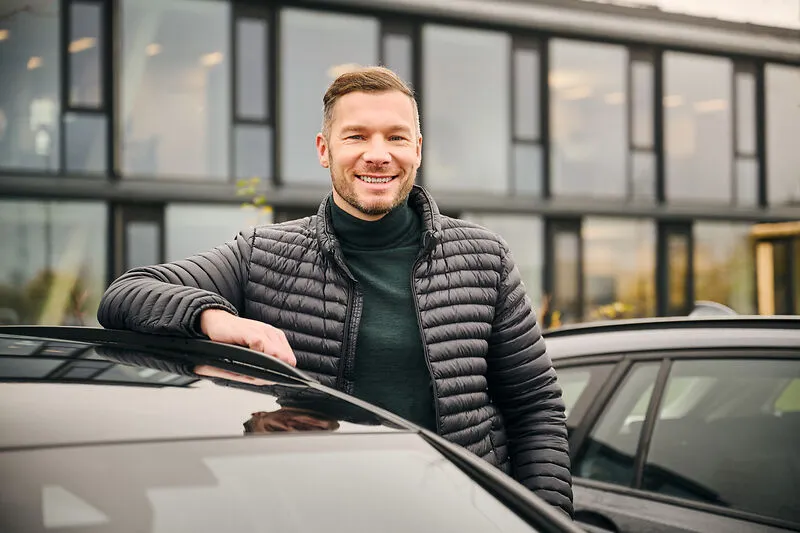 Ein lächelnder Mann lehnt an einem Auto vor dem modernen Bürogebäude von Liftstar GmbH im Gesundheitsbereich.