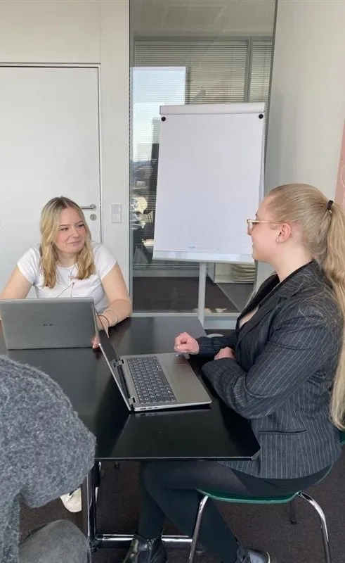 Drei junge Auszubildene Bürokaufleute bei Liftstar im Meeting, eine Person präsentiert am Flipchart, zwei hören zu und arbeiten am Laptop. Köln