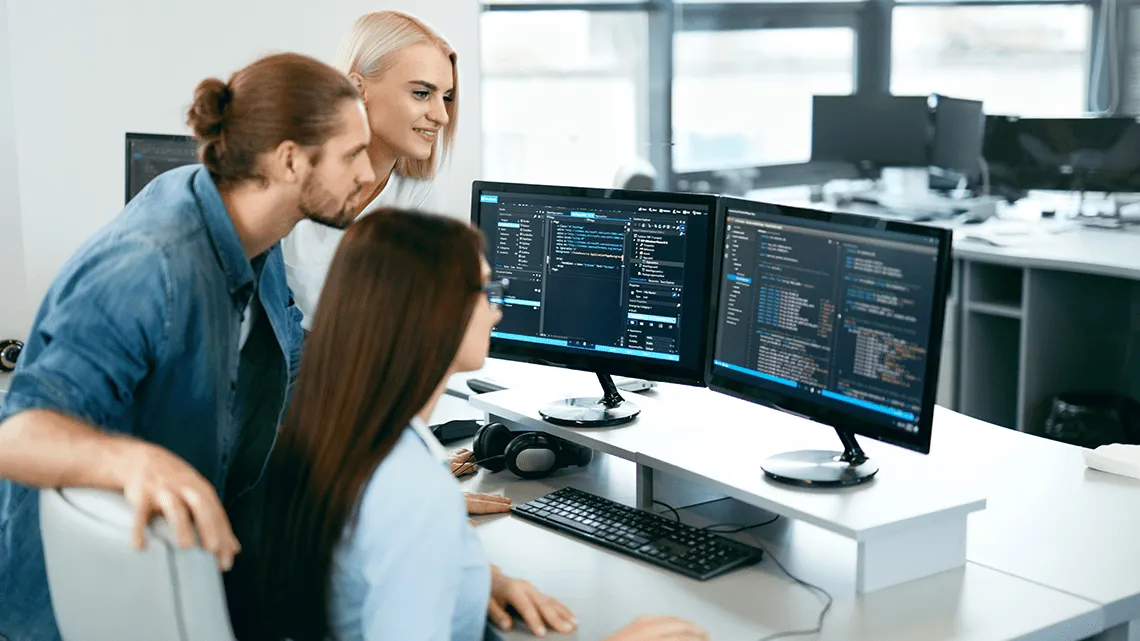 Drei IT-Kolleg:innen arbeiten gemeinsam an Code auf Monitoren in einem modernen Büro bei Logicalis GmbH.
