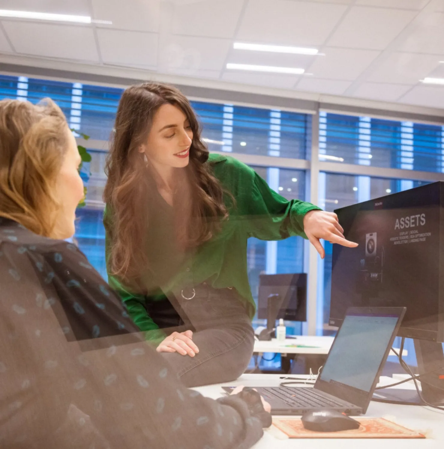 Two colleagues chatting at a desk while one points to a screen during a casual team meeting at Miele.
