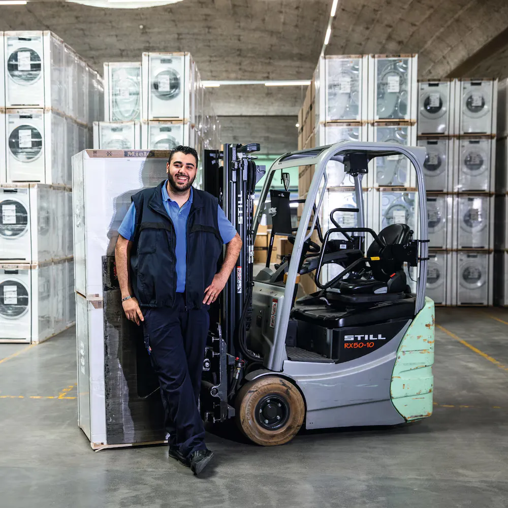 Ein freundlich lächelnder Miele-Mitarbeiter steht vor einem Gablestapler, auf dem zwei Miele Waschmaschinen geladen sind