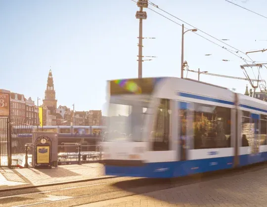 A tram speeds through sunlit Amsterdam street, with historic buildings in the background. Perfect for those who love the hustle and bustle!