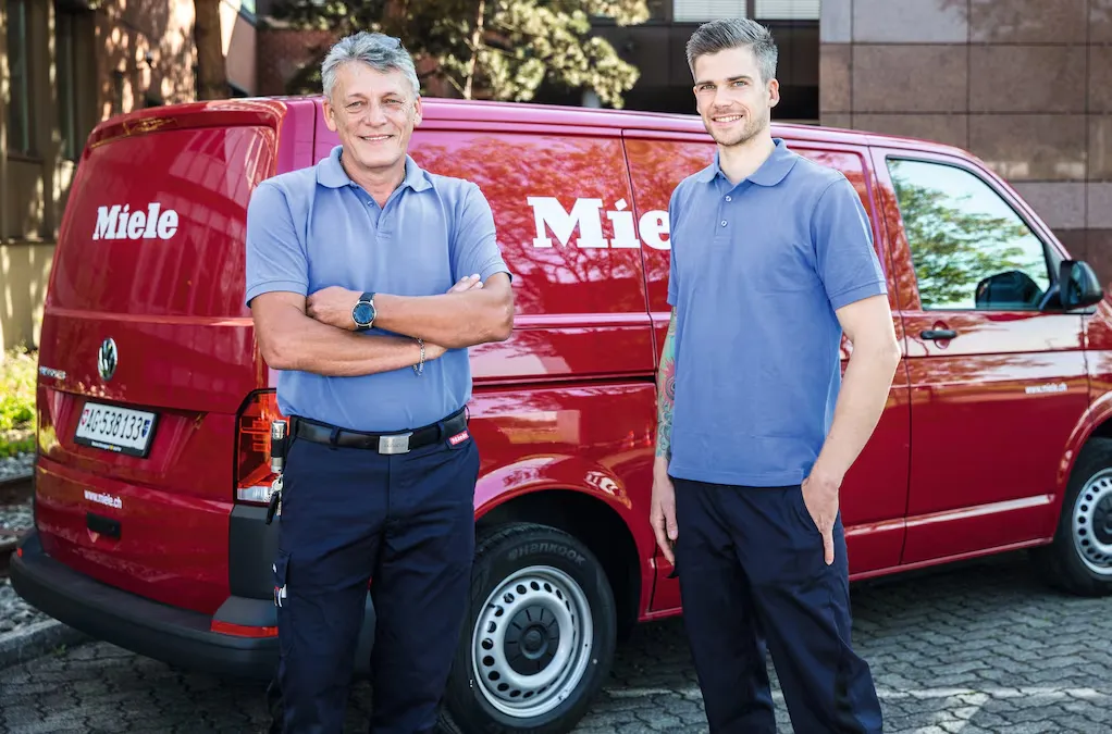 Zwei Miele-Mitarbeiter in blauen Polohemden stehen lächelnd vor einem roten Miele-Firmenwagen.