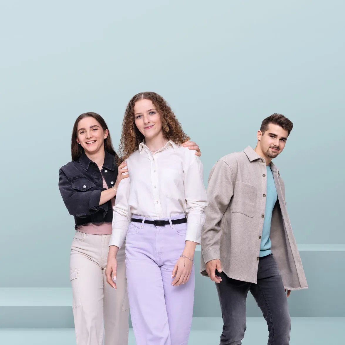 Three young people wearing casual clothing smiling at the camera against a light blue background.
