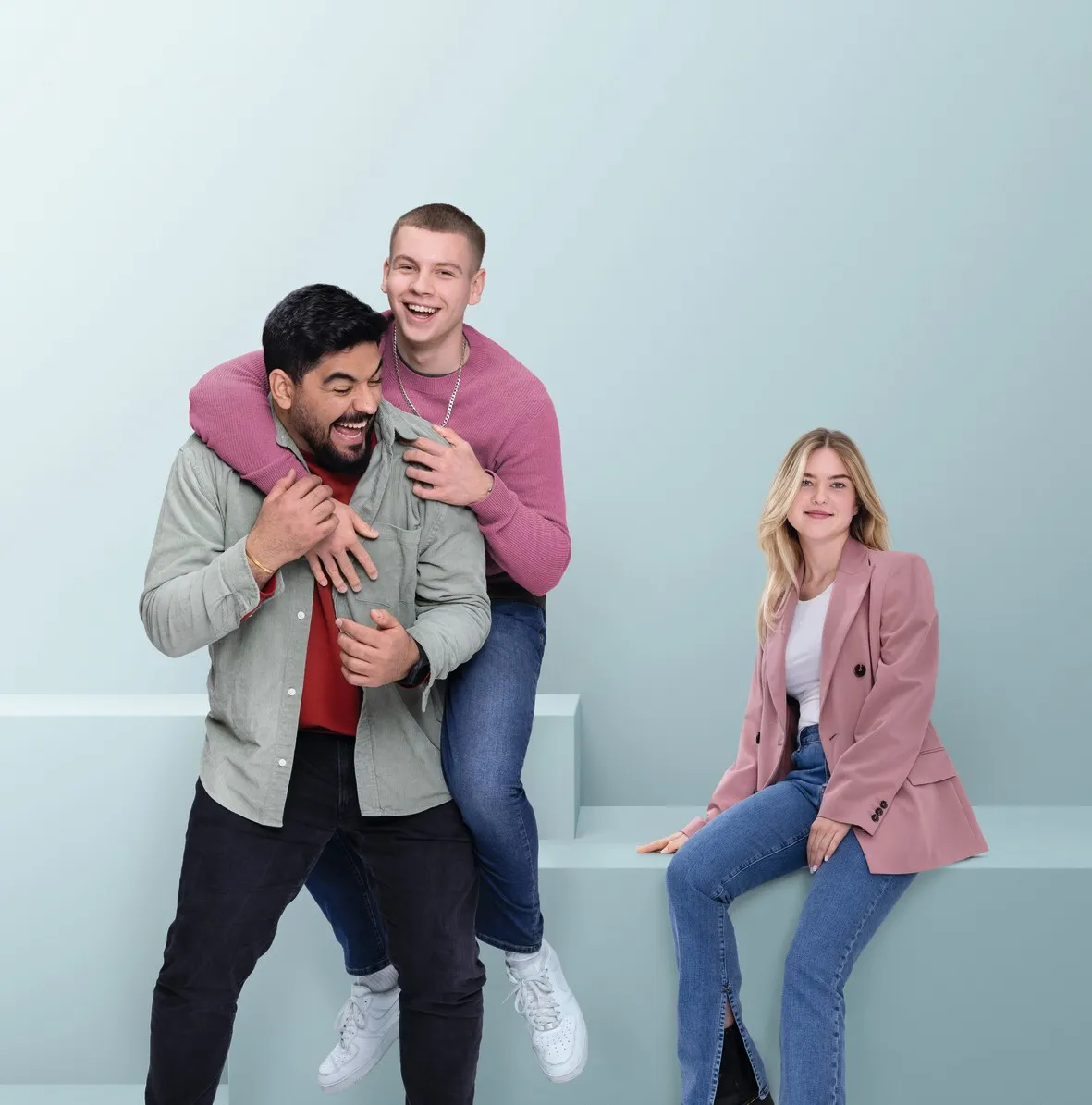 Drei junge Erwachsene lächeln in die Kamera, einer steht im Vordergrund, zwei sitzen und stehen im Hintergrund vor einem hellen Hintergrund.