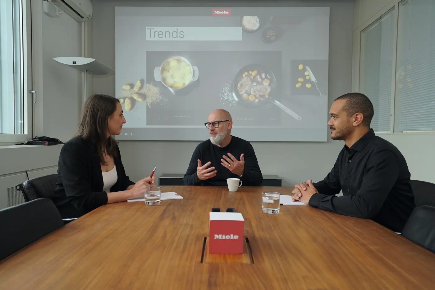Drei Personen in einem Meetingraum bei Miele, diskutieren vor einer Präsentation mit dem Titel "Trends".