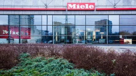 Budynek Miele Polska z przeszklonymi oknami, odbijający miejską panoramę, z czerwonym logo firmy na fasadzie.