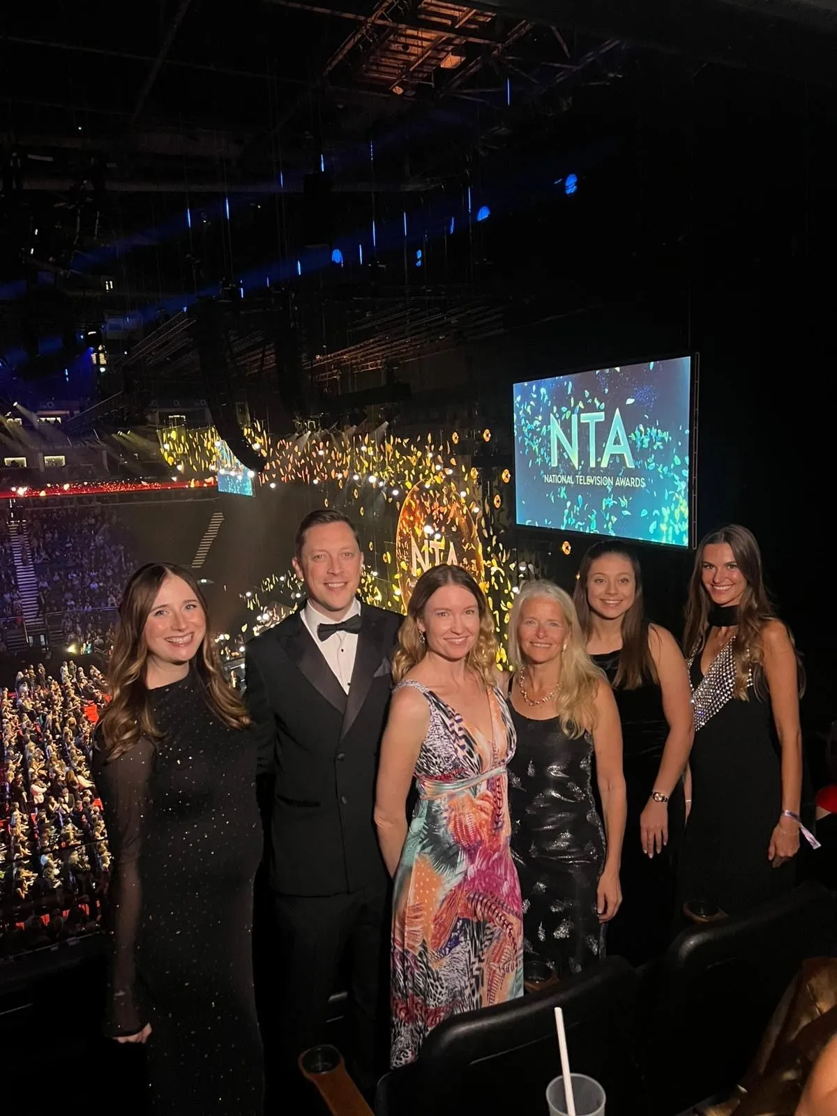 A group of six people dressed in formal attire, smiling at an award ceremony with "National Television Awards" displayed on a screen in the background.