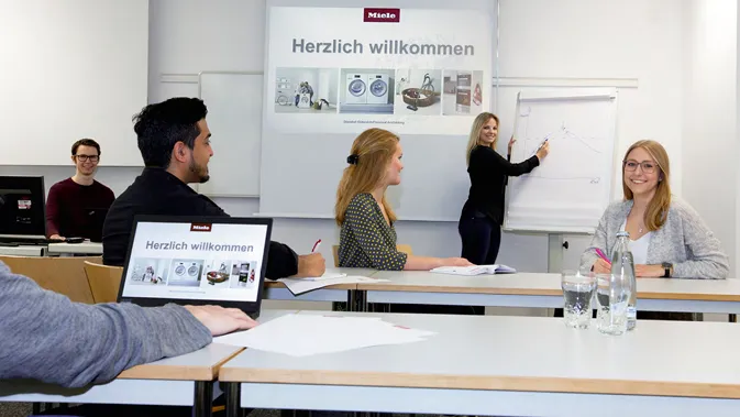 Un team di giovani professionisti discute in aula presso Miele, con una presentazione su schermo e lavagna a fogli mobili.