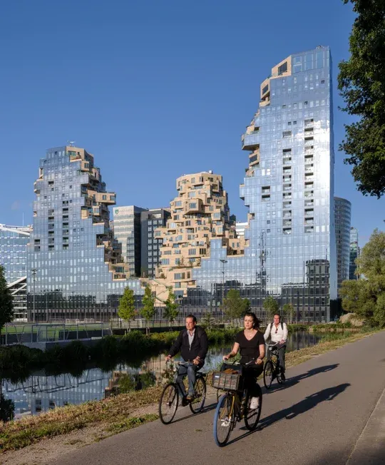 Three people cycling past a stunning, modern building with a unique, jagged design reflecting the blue sky.