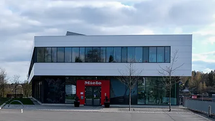 Modern Miele building in Norway with large windows and a red entrance, set against a cloudy sky, showcasing the brand's innovative spirit.