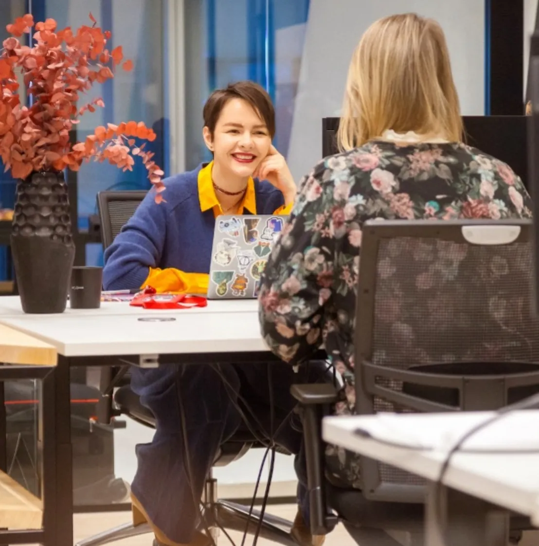Two colleagues chatting and smiling at a desk in a bright, modern Miele office, with a laptop and coffee nearby.