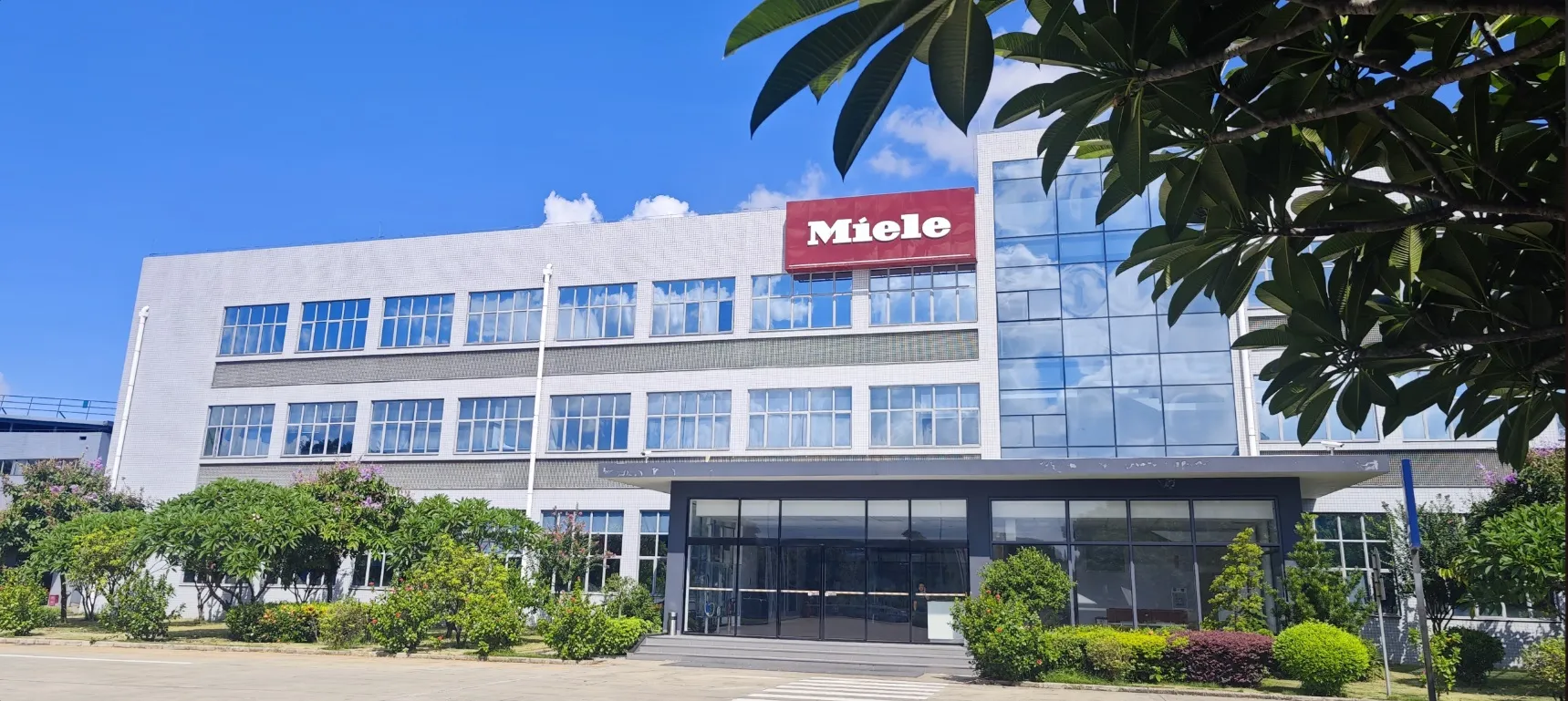 Außenansicht des Miele-Gebäudes von der Produktion in Dongguan mit blühenden Bäumen im Vordergrund und klarem Himmel.