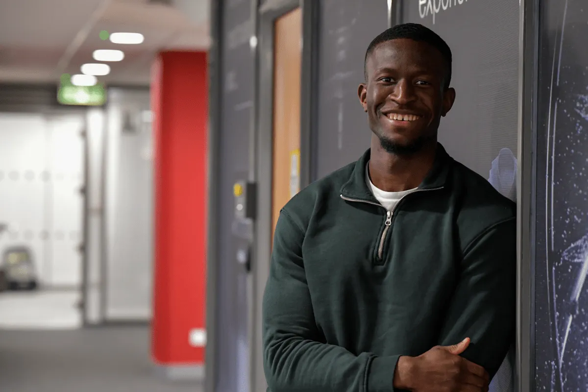 Tobi, Category Manager, smiling in a casual green sweater leaning against a wall at Miele & Cie. KG office, showcasing a friendly work environment.