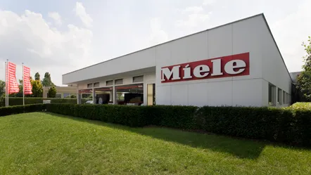 Het moderne Miele Luxemburg kantoorgebouw met bedrijfsnaam en vlaggen, omringd door groen.
