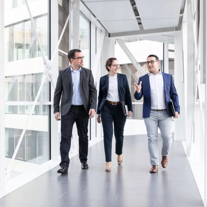 Drei Fachleute in Businesskleidung gehen lachend durch einen modernen Büroraum bei Miele & Cie. KG.