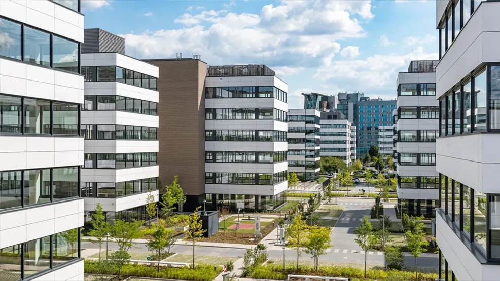 Nowoczesny kompleks biurowy Miele Global Services otoczony zielenią, z widokiem na kilka budynków w słoneczny dzień.