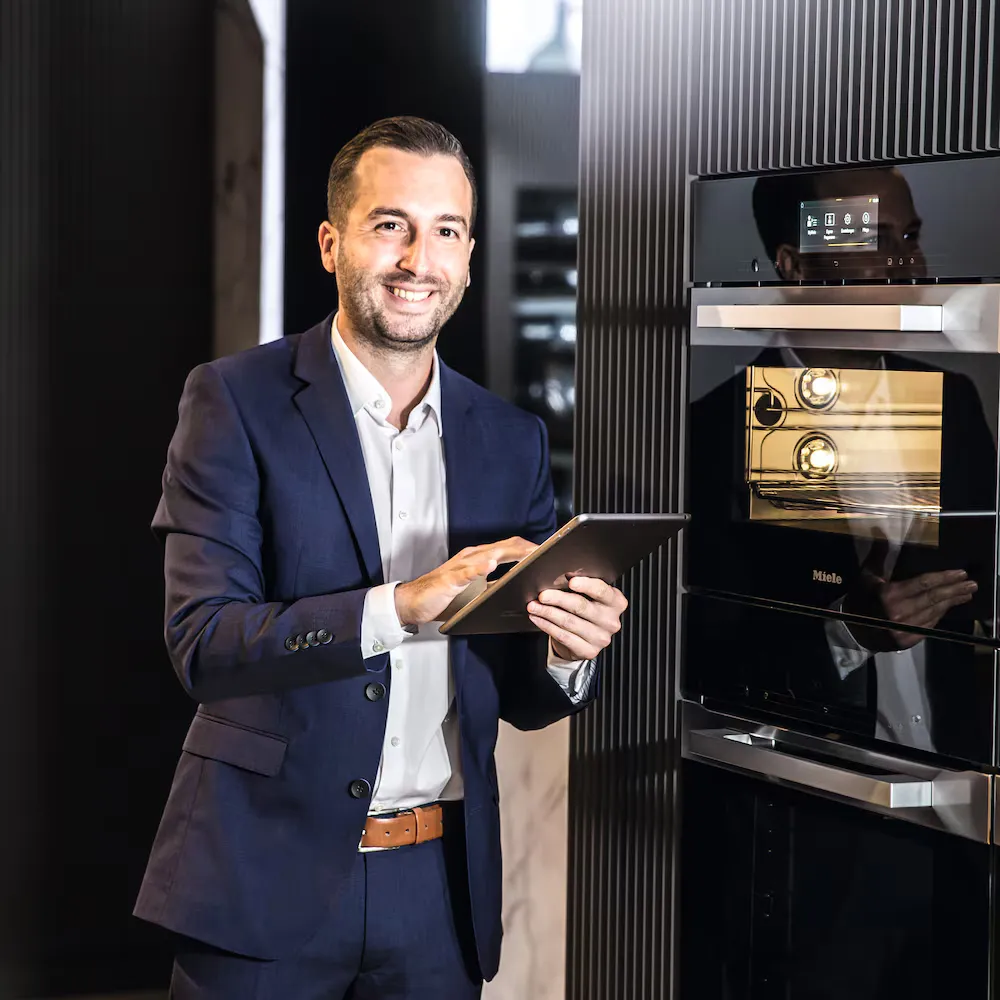 Ein freundlich lächelnder Sales-Mitarbeiter von Miele steht mit einem Tablet in der Hand vor einem modernen Backofen
