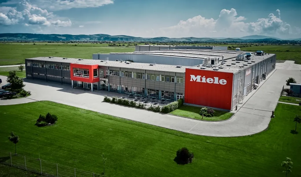 Hală modernă Miele într-un peisaj rural, cu cer senin și munți la orizont.